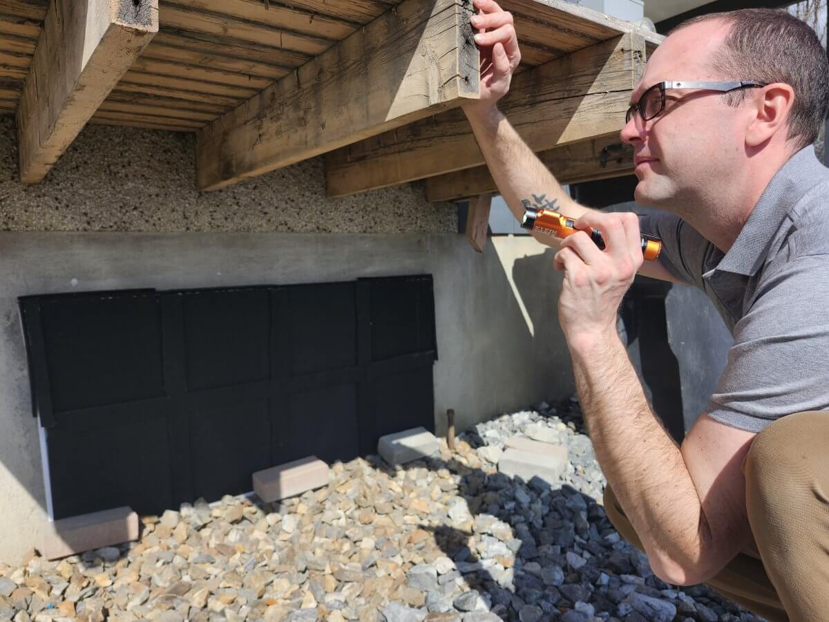 Inspector Evaluating a Deck and its Structure Erich from At Ease Property Inspections performs a detailed inspection, kneeling down to shine a flashlight on the rough-sawn wood support beams beneath an exterior deck, checking for structural integrity and damage. This type of thorough assessment is critical for buyers in Okanagan cities like Vernon and Armstrong, where properties often have outdoor living spaces.