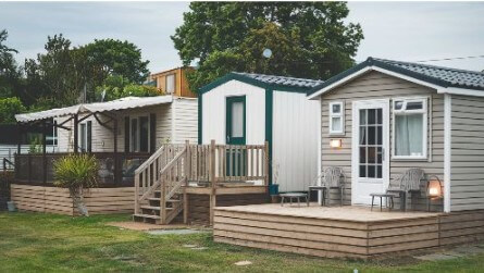 A photo of three houses on a property. The first house is a mobile home with a deck and a few plants. The second house is a modular home with a wooden staircase and a green door. The third house is a park model home with a few chairs on the deck and a lamp next to the door. The background contains trees and a fence. This is found on a dedicated home inspector website At Ease property Inspections