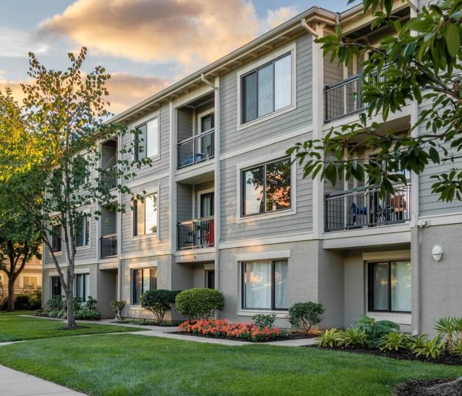 This is a normal bog-standard condo. This is a very standard style of condo building. A photograph of a three-story condo building with a modern architectural design and light gray exterior walls. Large windows are visible on each floor, reflecting the bright blue sky and surrounding greenery. A neatly manicured lawn stretches out in front of the building, complemented by a few mature trees and colorful flowerbeds. Warm sunlight gently illuminates the building, creating soft shadows and highlighting its clean lines. Inspector Erich Kamprath with At Ease property Inspections can inspect condos like this in Kamloops, Vernon, Lake Country, Kelowna, and more areas!