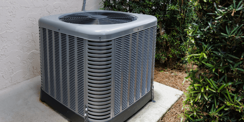 Outdoor air conditioning unit installed beside a residential home, surrounded by gravel and landscaping. The unit is part of a central cooling system, featuring a visible fan, metal housing, and ventilation grilles. It plays a crucial role in maintaining indoor comfort by releasing heat from inside the house. Regular maintenance of this AC condenser unit is essential for optimal performance, energy efficiency, and long-term reliability during warmer months. Erich Kamprath with At Ease Property Inspections always tests AC units to confirm they are working as Okanagan summers are very hot.