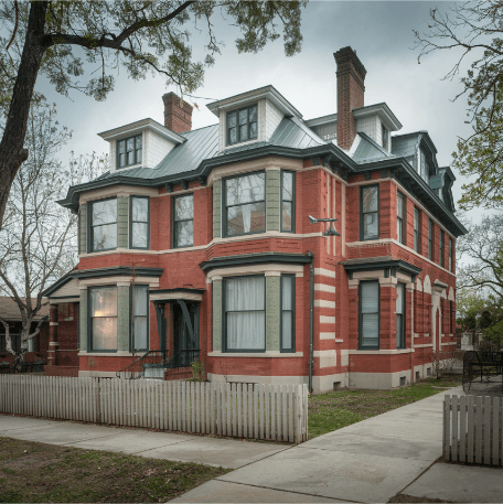 Brick Heritage Home A classic example of an early 20th century home with a red brick facade and a green roof. The house has multiple stories, each with large windows. There are two chimneys on the roof. The house is surrounded by a white picket fence and has a concrete path leading to the front door. There are trees in the front yard. The sky is overcast. Erich with at ease property inspection has inspected old homes just like this numerous times in Kamloops, Kelowna, Vernon, Okanagan or other nearby areas
