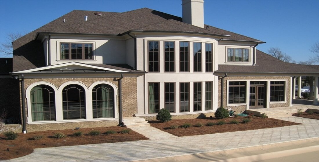 This image showcases a large, modern two-story residential home featuring a combination of brick and stucco siding, complete with a dark asphalt shingle roof and extensive window installations. The front facade highlights a variety of window types, including multiple tall, narrow fixed-pane windows, an elegant set of three arched windows, and several large double-hung and picture windows. From a home inspector's perspective, key elements to evaluate include the window seals, proper caulking around the frames, presence of weep holes for drainage, and overall alignment within the wall assembly. The home appears to have ample natural lighting, which is beneficial for energy efficiency but also warrants an inspection of potential UV damage to interior finishes. The brick veneer and stucco interfaces should be checked for signs of moisture intrusion or cracking. Inspectors would also note roof flashing above window bays and verify proper water runoff to prevent damage. The front landscaping and grading slope slightly away from the structure, a positive detail in preventing foundation water issues. This photo is ideal for illustrating topics such as window installation quality, exterior inspection points, and identifying potential maintenance areas in upscale residential construction. Erich Kamprath with At Ease Property Inspection is experienced and confident inspecting all window styles and home builds